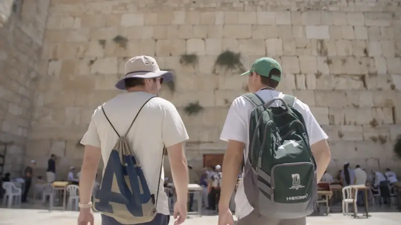 Two men with backpacks and hats walk towards the Western Wall in Jerusalem, with people gathered in the courtyard.